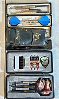 Two dart sets in their respective cases, showing the individual darts, flights, and accessories including soft tips, conversion points, and a pin or badge.