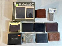 Lot of 9 leather wallets arranged in two rows. Top left is Timberland wallet in original packaging, bottom left is Champs wallet in box. Several used wallets in brown and black tones are displayed.