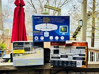 Three boxes of different motion activated light setups stacked on a wooden railing outdoors with trees in background.