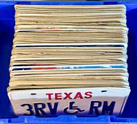 Blue bin filled with stacked license plates, front showing Texas disability plate '3RV RM'.