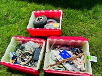 Three red and white bins outdoors on grass filled with electrical parts, bicycle components, and bicycle wheels
