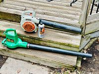 Two gardening blowers, one white and orange Stihl BG 55 with attached black tube, and one green electric Weed Eater Barracuda blower, shown together on outdoor wooden steps along with a can of MotoMix 50:1 fuel mixture.