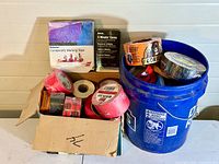 Wide view showing blue 5-gallon bucket filled with tape rolls and small boxes, plus cardboard box with tape rolls