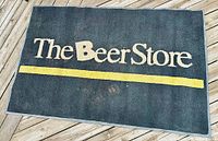 Front view of the black door mat showing 'The Beer Store' text and yellow line, some signs of wear.