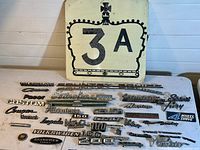 Photo of all vintage car badges arranged on a surface along with the Highway 3A road sign leaning behind them.