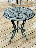 Full view of round cast iron table with ornate floral and leaf openwork tabletop, tripod base with curved legs and decorative braces, placed on wooden deck.