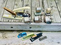 Photo showing entire lot of 11 vintage staplers and pencil sharpeners displayed on wooden steps. Top row includes metal pencil sharpeners with crank handles and larger staplers; bottom row has smaller staplers in various colors.