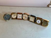 Seven vintage alarm clocks lined up on a flat surface showing full frontal faces and various shapes and sizes.