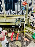Three string trimmers leaning against a deck railing: one gas trimmer and two corded electric models
