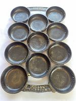 Top-down view of black cast iron poffertjes pan showing all 11 round cups arranged in a grid, with handle marked by raised text.