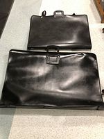 Two black artist portfolio cases lying flat; one larger with some dents, one smaller. Both have handles and shoulder straps attached.