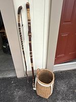 Photo of two hand carved walking sticks leaning against wall next to woven basket on ground.