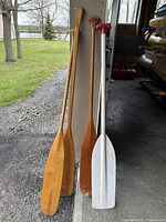 Four boat oars standing upright: three wooden oars and one white polymer oar with red handle.