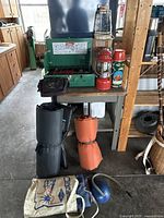 Green Coleman Easi-Lite camp stove, red and white Coleman lanterns, thermoses on a utility table with camping mattresses and bag underneath