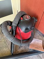 Photo showing the red and black Craftsman Shop Vac with flexible hose and instruction manual beside it.