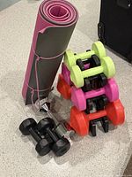 Photo shows two rolled yoga mats secured with pink straps, six pairs of dumbbells in various colors (black, chrome, lime green, pink, and orange) stacked on a black dumbbell rack on light gray flooring.