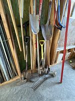 Photo showing various garden tools including shovels, rakes, and hoes leaning against a wood wall in a garage.