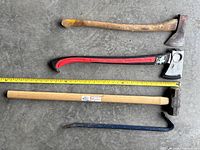 Full view of all tools laid on concrete floor with yellow tape measure for scale. Shows three axes, sledgehammer, and crowbar with worn wooden and fiberglass handles.