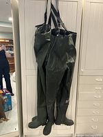 Two pairs of green rubber hip waders with boots and shoulder straps hanging side-by-side.