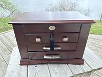 Front view of the closed dark wood jewellery box chest showing drawer fronts with silver-tone handles and lock with tassel key.