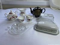 All items in the lot arranged on white background, showing Royal Albert cream and sugar set, Gibson & Sons black and gold teapot, two butter dishes, a glass juicer, and Royal Adderley cream and sugar set.