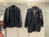Two black leather coats hanging side by side on hangers showing front view, one bomber style, the other medium length coat.