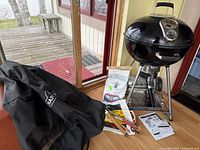 Overview of grill on tripod legs with included cover, charcoal bag, tools and manual