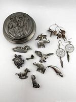 Photo showing closed pewter lidded trinket box featuring embossing of iris flowers, alongside animal-shaped pewter pins and two pairs of earrings including feather/dreamcatcher style