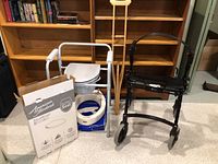 Photo of wooden crutches, white metal commode chair, mountable raised toilet seat, box for Apia AquaWash bidet, and Dana Douglas Handi-House rollator/walker.