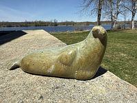 Side and top view of the greenish soapstone sea lion carving placed outdoors on a concrete ledge with a river and trees in background.
