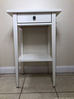 Front view of white wooden side table showing drawer, lower shelf, and legs against a wall and tiled floor.
