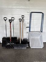 Full view of 7 black plastic snow shovels and 2 large snow scoops, various handle materials including wood, metal, and plastic grips, showing wear and some paint chipping.