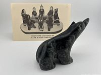 Profile view of the dark soapstone bear sculpture with a card showing other soapstone carvings by Mark & Ross Parkinson behind it.