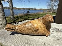 Full side view of the soapstone sea lion carving showing its textured surface and natural coloration with the sea and trees in background.