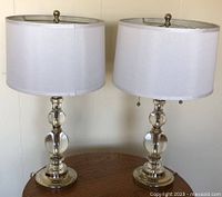 Front view of two matching glass and chrome table lamps on a wooden round table, showing white fabric drum shades and double glass ball bases with chrome accents