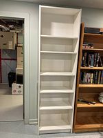 Front view of the white IKEA bookshelf placed indoors near another wooden bookshelf, showing all five adjustable shelves.
