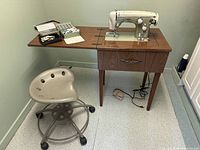 Sewing machine placed on a wooden cabinet stand with a beige metal round stool in front on casters. Accessories including foot pedal and small boxes on top of stand.