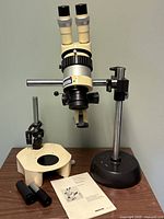 Microscope head assembly mounted on rod stand and stage base with eyepieces and manual on tabletop