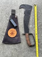 Full view of Woodman's Pal utility hatchet next to measuring tape with black fabric sheath nearby, hatchet has a hooked blade and leather wrapped wooden handle