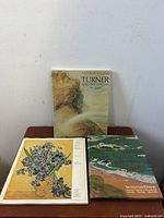 Three vintage art books on a wood surface, seen from front and top angles, showing titles and covers