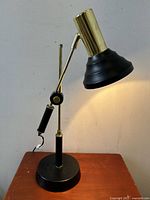 Side view of the adjustable industrial style pivoting desk lamp showing black conical shade, brass pivot arm, and circular base on wooden surface.