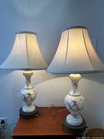 Pair of antique enameled glass table lamps with beige fabric shades on a wooden table, shown plugged in and switched on.