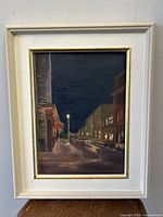 Frontal view of the framed nighttime street scene oil painting showing illuminated storefronts and street lamps with reflections on the pavement.