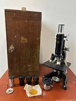 Complete microscope and accessories displayed on wooden surface with oak case in background.