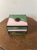Front view of the small cube-shaped stained glass box with pink and green panels and red stone on lid.