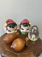 Front view of the full set showing two ceramic Lefton Pixie Elf salt and pepper shakers with red hats and green collars, two teak fish salt and pepper shakers, and a clear Lucite timer with sand inside, all placed on a wooden surface.