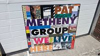 Full view of the sealed Pat Metheny Group "We Live Here" poster laid against a wall showing title and partial signatures.