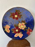 Front view of the plate leaning against a beige wall showing the full floral design with various colored roses on a blue textured background.