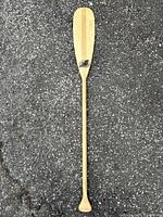Full view of the wooden canoe paddle laid flat on asphalt surface, showing the entire length and shape from grip to blade.