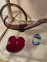 Glass paperweight, elongated purple glass bowl and red ashtray displayed on glass table.